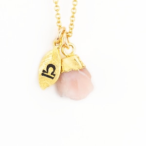 May include: Gold-colored necklace with a Libra zodiac charm and a pink gemstone pendant. The Libra charm is a gold-colored leaf with a black Libra symbol. The gemstone is a light pink color.