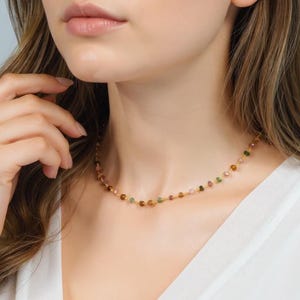 May include: A delicate gold necklace featuring small, colorful gemstone beads. The necklace has a simple design with a strand of round beads in shades of pink, green, and brown. The necklace is worn around the neck.
