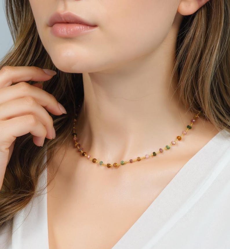 May include: A delicate gold necklace featuring small, colorful gemstone beads. The necklace has a simple design with a strand of round beads in shades of pink, green, and brown. The necklace is worn around the neck.