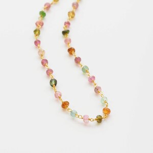 May include: A delicate necklace featuring small, multi-colored gemstone beads strung on a gold-colored chain. The gemstones are in shades of pink, green, orange, and yellow. The necklace is displayed against a white background.