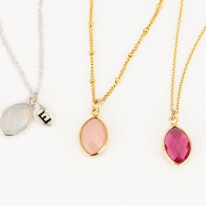May include: Three necklaces featuring marquise-cut pendants. The first is a silver chain with a translucent white stone and a small oval charm stamped with the letter E. The second is a gold chain with a pale pink stone. The third is a gold chain with a vibrant fuchsia stone.