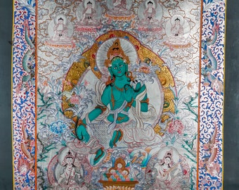 May include: A traditional Tibetan thangka painting depicting a green deity seated on a lotus throne. The deity is surrounded by smaller figures and intricate floral patterns. The painting is framed in a blue and gold border with a Thankka.com watermark.