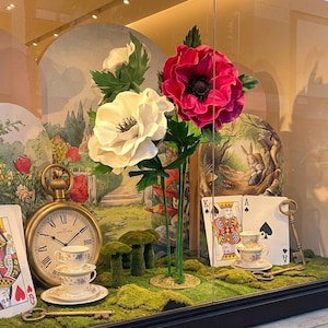 May include: A whimsical window display featuring oversized paper flowers in white and magenta, playing cards, a pocket watch, and miniature teacups. The display has an Alice in Wonderland theme, with a backdrop of a garden scene and a key.