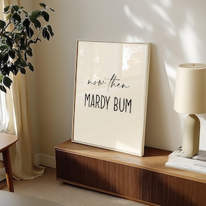 May include: A framed art print with the words "now then MARDY BUM" in black script and block letters. The print has a cream-colored background and a thin, gold-colored frame. The artwork is displayed on a wooden cabinet.