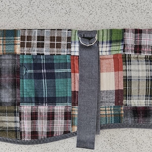 May include: A patchwork dog coat featuring a variety of plaid patterns in shades of red, green, blue, and brown. The coat has a gray fleece collar and a gray strap with a metal ring. The initials "ERH" are embroidered on a patch.