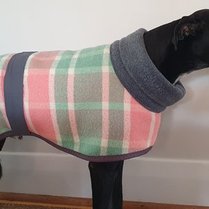 May include: A black dog wearing a dog coat with a pink, green, and grey plaid pattern. The coat has a grey fleece collar and a dark blue band down the back. The dog is indoors.