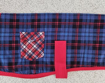 May include: A dog coat with a blue, black, and red plaid pattern. The coat has a red fleece collar and trim, a red plaid pocket, and a red strap. A small label is sewn on the lower right side.