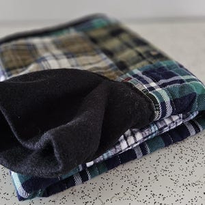 May include: Folded plaid flannel shirt with a black fleece lining. The shirt features a green, blue, white, and brown plaid pattern. The black fleece lining is visible at the cuff. The shirt is folded neatly.