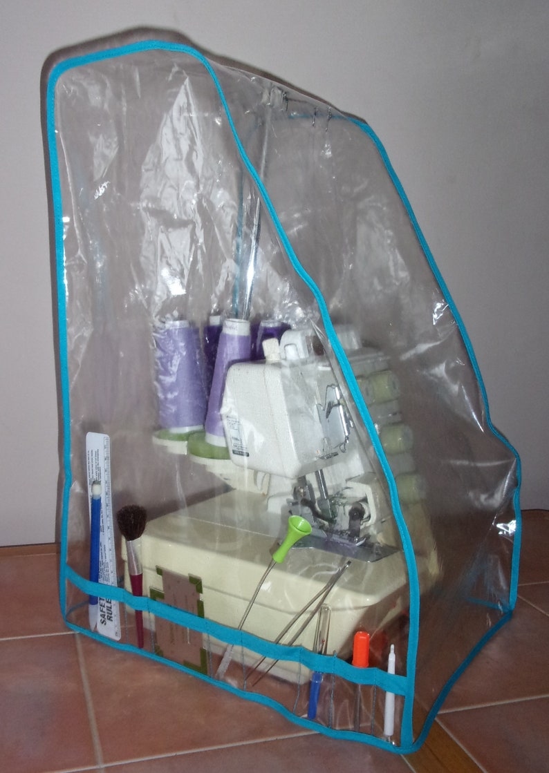 May include: A clear plastic cover with a turquoise trim protects a white sewing machine with spools of thread and various tools. The cover has pockets for storing sewing supplies. The text "Safety Rule" is visible on the cover.