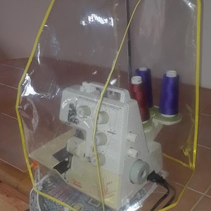 May include: A white sewing machine with a clear plastic cover with a yellow trim. The machine is on a table with a patterned fabric hanging from the side. There are spools of thread in the machine.
