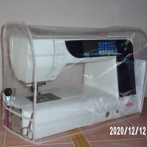 May include: A white sewing machine with a clear plastic cover. The machine has a digital display and a needle threader. The machine is sitting on a tiled surface.