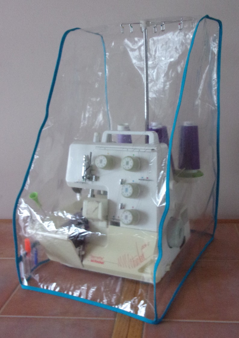 May include: A clear plastic cover with a turquoise trim protects a white sewing machine with multiple dials and spools of thread.