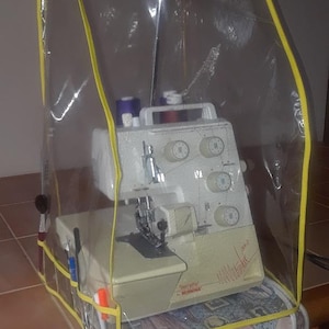 May include: A white sewing machine with a clear plastic cover with yellow trim. The machine is sitting on a table with a floral fabric underneath.
