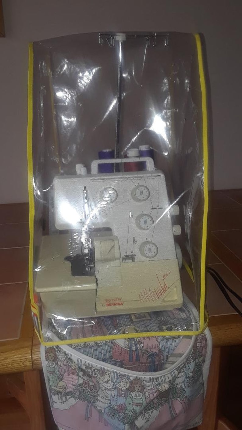 May include: A white sewing machine with a clear plastic cover and a yellow trim. The machine is sitting on a table with a pink and white fabric bag underneath.