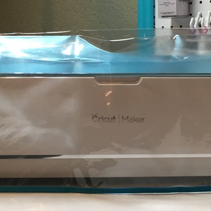 May include: A clear plastic cover with a turquoise trim protects a white Cricut Maker cutting machine.