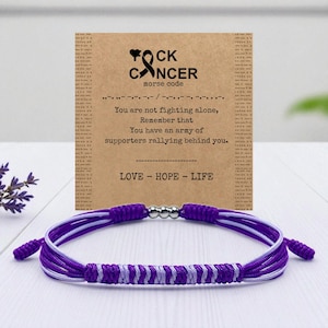 May include: A purple and white braided bracelet with a silver clasp is shown in front of a card. The card has the words "FOCK CANCER" and "LOVE - HOPE - LIFE" printed on it, along with a message about support.