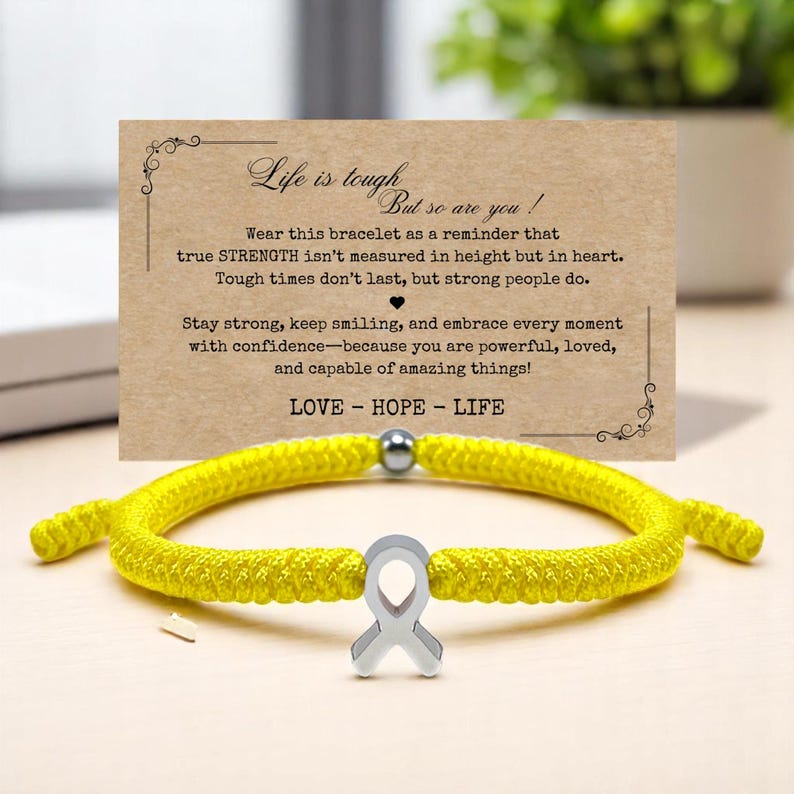 Peut inclure: Un bracelet en corde jaune avec un charm en forme de ruban argent&eacute;. Une carte derri&egrave;re le bracelet indique "Life is tough but so are you !" et comprend les mots "LOVE - HOPE - LIFE". Le bracelet est sur une surface claire.