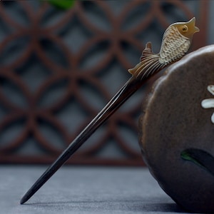 May include: A brown and white hair stick with a carved fish design. The stick is made of horn and has a smooth, polished finish.