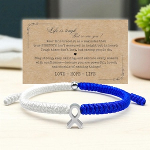 May include: A white and blue braided bracelet with a silver ribbon charm. The bracelet is displayed in front of a card with the text "Life is tough, but so are you!" and inspirational messages. The bracelet is a symbol of hope.