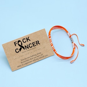 Op de afbeelding: Oranje en wit touw armband met een zilveren kraal. Het armband ligt op een bruine kaart met de tekst "FOCK CANCER" en "morse code armband". De kaart bevat ook de boodschap "Je vecht niet alleen. Onthoud dat je een leger van supporters hebt die achter je staan."