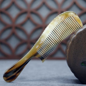May include: A brown and tan comb made of horn with a curved handle. The comb has fine teeth and is resting on a brown surface.