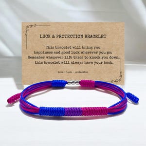 May include: A blue, purple, and pink braided bracelet with a silver charm. The bracelet is on a white surface with a card that reads "LUCK & PROTECTION BRACELET" and a message about the bracelet bringing happiness and good luck.
