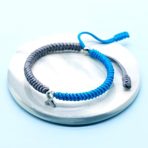 May include: A two-tone bracelet with a blue and gray braided design. The bracelet features a silver-colored charm and adjustable blue cord ends. The bracelet is displayed on a white, circular surface against a light blue background.