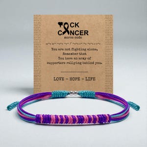 Pode incluir: Uma pulseira colorida com uma mensagem de apoio. Apresenta tons de roxo, rosa e turquesa, com um fecho prateado. Um cartão mostra "F*ck Cancer" em código Morse, com as palavras "Amor, Esperança, Vida".