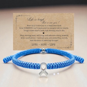 May include: A blue braided bracelet with a silver ribbon charm and a small silver bead. A card behind the bracelet reads "Life is tough, but so are you!" with inspirational text about strength and love.
