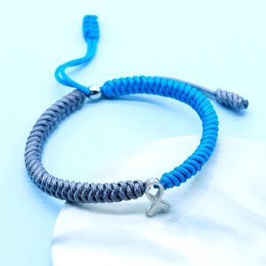 May include: A blue and gray braided bracelet with a silver ribbon charm. The bracelet is adjustable with a sliding knot closure. The charm is a small, silver ribbon. The bracelet is on a light blue background.