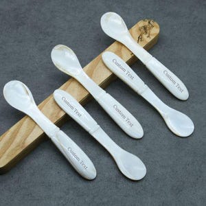 May include: Five white mother of pearl spoons with a rounded bowl and a long handle. Each spoon has the text "Custom Text" engraved on the handle.