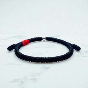 May include: Black braided bracelet with a red accent and a silver-colored bead. The bracelet is made of a thick, woven material and is designed to be worn around the wrist. The bracelet is shown against a white background.