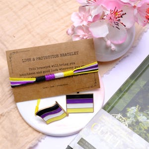 May include: A non-binary pride bracelet with yellow, white, purple, and black threads, alongside two enamel pins depicting the non-binary flag. A card reads "LUCK & PROTECTION BRACELET". The items are on a white plate.