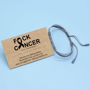 Pode incluir: Pulseira tecida cinza com uma fita preta e o texto "FOCK CANCER" e "pulseira de código morse". O texto abaixo diz "Você não está lutando sozinho. Lembre-se de que você tem um exército de apoiadores que o apoiam."