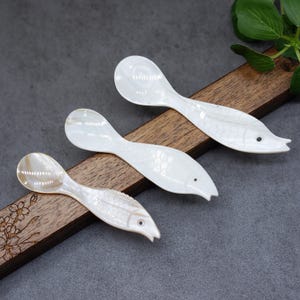 Könnte beinhalten: Drei weiße Perlmuttlöffel in Fischform. Die Löffel haben eine kleine, abgerundete Schale und einen langen, schlanken Stiel, der an der Spitze spitz zuläuft. Die Schuppen des Fisches sind in den Stiel geschnitzt.
