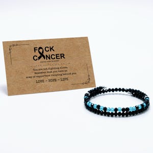 May include: A black beaded bracelet with light blue beads, displayed with a brown card. The card reads "FOCK CANCER" and the words "LOVE - HOPE - LIFE". The bracelet is a symbol of support.