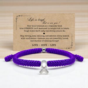 May include: A purple braided bracelet with a silver ribbon charm. The bracelet is displayed in front of a card with the text "Life is tough, but so are you!" and the words "LOVE - HOPE - LIFE".