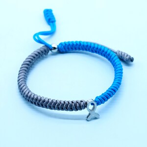 May include: A blue and gray braided bracelet with a silver ribbon charm. The adjustable bracelet features a sliding knot closure and is displayed on a light blue background. The bracelet is a stylish accessory.
