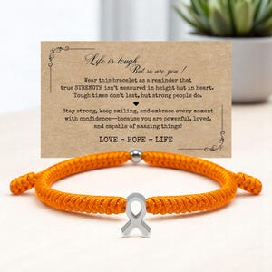 Peut inclure: Bracelet en cordon orange avec un charm en forme de ruban argent&eacute;. Le bracelet est pr&eacute;sent&eacute; devant une carte avec le texte "Life is tough, but so are you !" et les mots "LOVE - HOPE - LIFE".