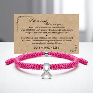 Peut inclure: Un bracelet tress&eacute; rose vif avec un charm en forme de ruban argent&eacute;. Le bracelet est pr&eacute;sent&eacute; devant une carte avec le texte "Life is tough, but so are you !" et un message inspirant.