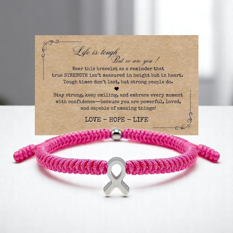 Peut inclure: Un bracelet tress&eacute; rose vif avec un charm en forme de ruban argent&eacute;. Le bracelet est pr&eacute;sent&eacute; devant une carte avec le texte "Life is tough, but so are you !" et un message inspirant.