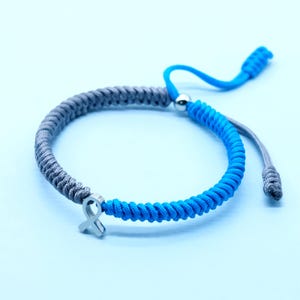 May include: A two-tone bracelet with a blue and gray braided cord design. A silver-colored ribbon charm is attached to the bracelet. The bracelet has an adjustable sliding knot closure. The background is a light blue color.