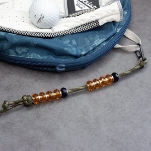 May include: A golf ball and glove rest inside a blue golf bag. A beaded golf score counter with amber and black beads hangs in front of the bag. The score counter is attached to a camouflage green cord.