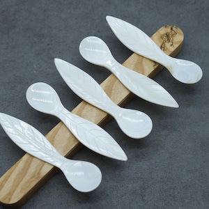May include: Six white mother of pearl spoons with leaf designs on a wooden surface. The spoons are arranged in a row, with the handles pointing to the right.