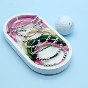 May include: A collection of colorful beaded bracelets in a white oval tray, with a golf ball in the background. The bracelets feature silver beads and various cord colors, including pink, black, green, and white.