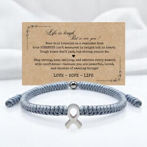 Peut inclure: Un bracelet tress&eacute; gris avec un charm en forme de ruban argent&eacute; et une petite perle argent&eacute;e. Une carte derri&egrave;re le bracelet indique "La vie est dure, mais vous aussi !" et comprend les mots "AMOUR - ESPOIR - VIE".