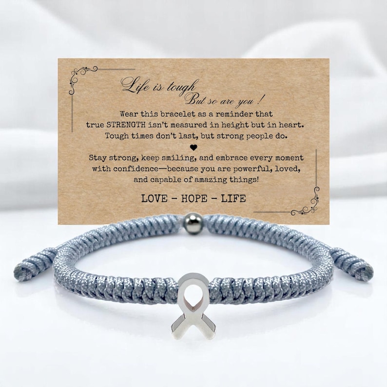 Peut inclure: Un bracelet tress&eacute; gris avec un charm en forme de ruban argent&eacute; et une petite perle argent&eacute;e. Une carte derri&egrave;re le bracelet indique "La vie est dure, mais vous aussi !" et comprend les mots "AMOUR - ESPOIR - VIE".