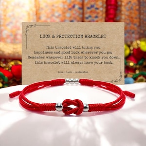 May include: Red string bracelet with silver beads and a knot in the centre. The bracelet is on a white surface with a card that reads "LUCK & PROTECTION BRACELET" and a message about the bracelet bringing happiness and good luck.