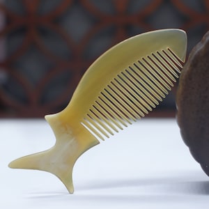 May include: A light brown comb shaped like a fish with a wide, flat handle and a row of fine teeth. The comb is made of natural horn and has a smooth, polished finish.