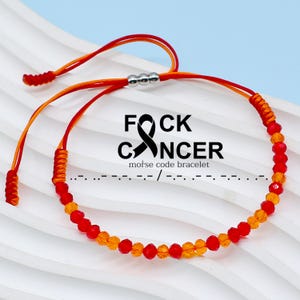 May include: Red and orange beaded bracelet with a silver charm and a red ribbon tied in a knot. The bracelet has the words "F*CK CANCER" and "morse code bracelet" written on it.